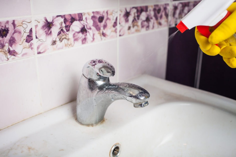 A person wearing yellow gloves sprays cleaner on a bathroom sink faucet with visible hard water stains.